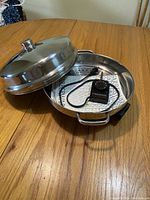Electric skillet on wooden table with lid placed beside it, showing inside steamer insert and heat controller device.