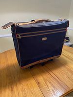 Front view of navy blue garment bag on wheels with brown leather trim, a zippered exterior pocket and top handle.