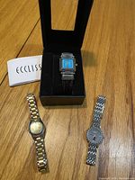 Three wristwatches on wooden surface: left is men's two-tone silver and gold watch, center is Ecclissi women's watch in black box with turquoise square face and black leather strap, right is silver tone women's wristwatch with black pinstriped round face and linked metal band.