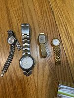 Four wristwatches laid out on wood floor, two silver-tone with metal link bands and two gold-tone with link bands, mixing round and square dials.
