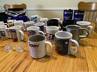 Photo showing assortment of mostly ceramic coffee mugs with designs, travel mugs, and glass shot glasses on wooden table