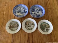 Five decorative plates on wooden table: two plates with blue and white winter barn scenes, three plates with multicolor winter scenes and gold rims.