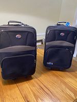 Front view of two black overnight luggage bags with oval logo, standing side by side on wooden floor under natural light.