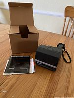 Polaroid Sun 600 LMS instant camera, original box, and instruction manual on wooden table with chair in the background