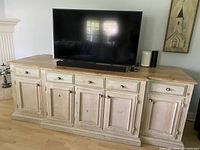 Front view of large solid wood entertainment console showing the oak top, antiqued white distressed finish with green drawer accents, five drawers, and five cabinet doors. Scratches visible on surface.