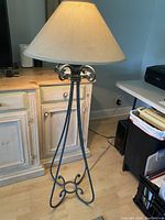 Full view of the tall floor lamp with Verdi green metal base and beige lampshade on a hardwood floor near a desk and cabinet.