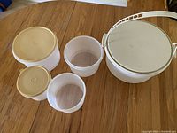 Overview of six white plastic Tupperware containers on wooden table with three smaller round containers having beige lids and three medium or small containers without lids along with a large white round cake container with a beige handle and lid.