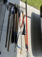 Photo of assorted garden tools including shovels, rakes, hedge shears, and other hand tools laid out on concrete with visible grass in background.