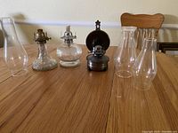 Three vintage hurricane lamps with glass bases and metal burners, plus separate glass chimneys placed on a wooden table.