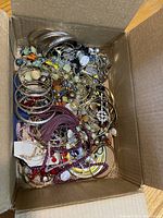 View of assorted costume jewelry pieces including metal bangles, beaded necklaces and earrings in a cardboard box.
