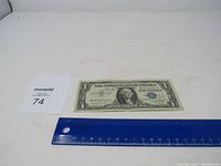 Front side of 1957 A Silver Certificate blue seal one dollar bill next to a ruler showing size for scale.