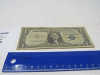 Front side of the 1957A Blue Seal $1 Silver Certificate with blue seal visible and an adjacent ruler showing the approximate length of the bill.