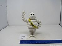 Front view of white cast iron Michelin Man bank with yellow Michelin sash and waving gesture, includes ruler for scale.