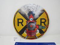 Front view of the round metal railroad sign with painted steam locomotive in front of traditional yellow and black crossbuck symbol. Minor rust and wear visible on edges.
