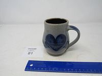 Front and angle views of a gray Rowe Pottery crock-style vase with blue heart and leaf designs and a handle, ruler included for size reference.