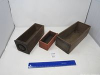 Three antique wooden boxes displayed with a ruler for scale, showing rectangular shapes and varying sizes and wood tones.