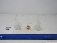 Three glass swan figurines including two large clear swans and one smaller orange-accented swan shown with ruler for scale.