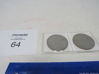 Two Eisenhower dollar coins in clear protective holders side by side with a ruler and number 64 card visible.