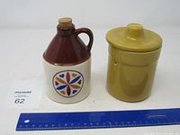 Photo showing a tan and brown ceramic jug with cork stopper and colorful star and heart pattern, and a mustard yellow lidded crock.