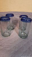 Four clear blown glass tumblers with blue rims arranged in a group on a quilted surface.