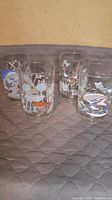 Four Disney-themed vintage 1996 anniversary glasses arranged on a quilted surface, showing overall wear and design of each glass.