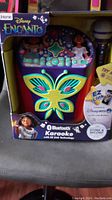 Front view of the Disney Encanto karaoke machine in box showing butterfly design and characters from the movie, microphone included on side.