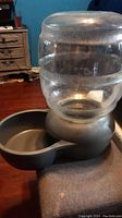 Clear plastic water container with condensation, placed on gray plastic base with water dispensing bowl showing wear and scratches.