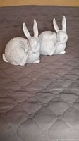 Photo showing two gray and white polyresin rabbit figurines curled and resting with ears upright on a quilted surface.