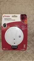 Front photo of Kidde smoke alarm in clear packaging with red backing card. Shows the round white smoke alarm and included 9V Eveready battery.