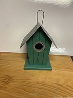 Front view of green wooden birdhouse showing circular entrance hole with metal reinforcement and metal hanging hook on roof peak.