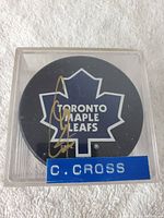 Top view of the autographed Toronto Maple Leafs hockey puck with Cory Cross signature visible inside a clear display case with a blue label reading 'C. CROSS'.