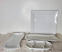 Seven white ceramic serving dishes including large square dish, various rectangular platters, rosebud dish, and small divided dish, displayed on a towel against white background.