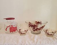 Front view showing one large Wheaton glass popcorn bowl, two small matching bowls, and a Catamount plastic popcorn popper with red graphics and lid.