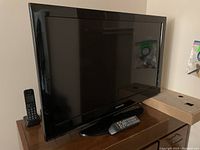 32 inch Samsung LCD TV placed on a wooden cabinet with two remote controls: one TV remote on the screen base and one cordless phone remote next to the TV. The TV is black with a glossy finish and powers on.