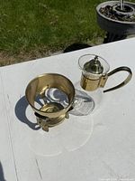 Glass jug with brass lid and handle along with brass-colored metal stand for holding the jug, photographed on white table outdoors with grass in background.