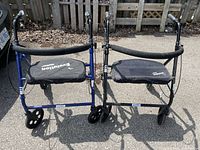 Photo of two Evolution handicapped walkers side by side outdoors on pavement in front of wooden fence.