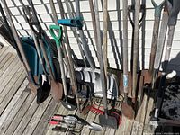 Photo showing an assortment of yard tools including shovels, snow shovels, rakes, hoes, and hand tools arranged on a wooden deck. Tools show wear and rust, used condition.