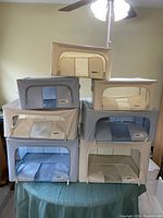 Seven fabric storage organizers in beige, cream, blue, and gray colors stacked on a table against an off-white wall with ceiling fan above.