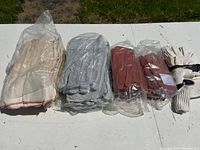 Five pairs of assorted gardening gloves laid out on a white surface against a grass background. Includes canvas, leather in gray and red colors, and knit gloves. Some gloves are sealed in plastic packaging.