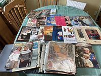 Photo showing a large selection of knitting and sewing related magazines, pattern books, and an open binder filled with pattern pages on a green table