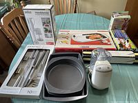 Braun handblender with chopper attachment box, Centro stainless steel flatware set box, several black non-stick baking pans, and a white electric small kitchen appliance on a green table.