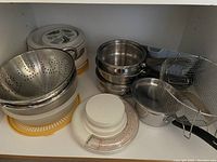 Photo showing a collection of assorted stainless steel pots and bowls stacked on the left side; two metal mesh strainers with handles on the right; and a white circular mechanical kitchen scale positioned in front.