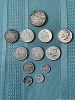 Top-down image showing twelve silver coins arranged on a blue fabric surface including Canadian and US coins of various denominations and years.