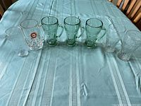 Nine assorted drinking glasses from Coca-Cola, Pepsi, and A&W arranged on a blue tablecloth: three green Coca-Cola mugs, one clear Pepsi mug with sticker, clear Coca-Cola glasses, and one clear A&W mug.