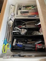 Photo of drawer full of various kitchen utensils, showing tools including scissors, graters, peelers, tongs, measuring spoons, and a garlic press.