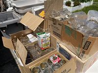 Multiple cardboard boxes filled with various sizes of clear Mason canning jars plus lots of loose lids and other small containers on an outdoor table.