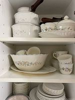 White ceramic kitchenware with floral designs arranged on shelves including pots with lids, bowls, cups and plates.