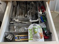 Image showing a kitchen drawer filled with assorted silverware including forks, spoons, knives and various kitchen utensils such as peelers, bottle openers, whisks, and new packaged item. The drawer organizer is a metal wire mesh tray divided into compartments.