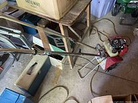 Photo of mini rototiller showing long metal handles, red body, and white fuel tank from a side angle.