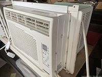 Side-angle view of NOMA white window air conditioner showing front air vents, control panel, and side model number label.